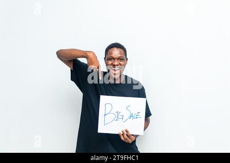 Aufgeregter afrikanischer Mann mit schwarzem Hemd, der auf eine weiße Karte mit Big Sale-Aufschrift zeigt und damit den Abschluss großer Verkäufe ankündigt. Black Friday s Stockfoto