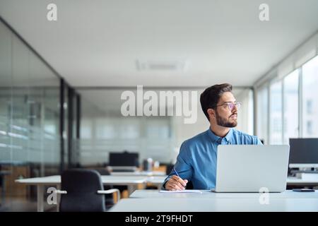 Geschäftiger lateinamerikanischer Geschäftsmann, der im Büro mit einem Laptop arbeitet, der wegblickt und denkt. Stockfoto