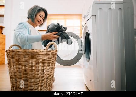 Eine glückliche Seniorin, eine lächelnde Großmutter, lädt schmutzige Kleidung in die Waschmaschine Stockfoto