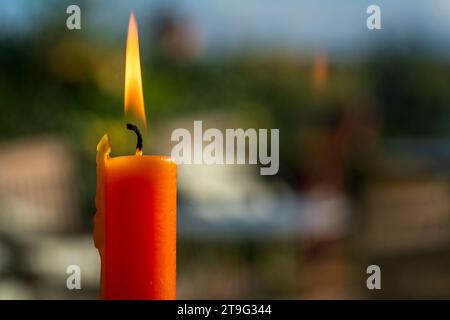 Eine einsame orangene Kerze strahlt ein warmes Leuchten in einem Fenster aus, die Flamme flackert vor einem verschwommenen Hintergrund im Freien. Die sanfte Beleuchtung erzeugt einen SER Stockfoto