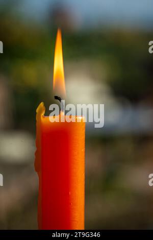 Eine einsame orangene Kerze strahlt ein warmes Leuchten in einem Fenster aus, die Flamme flackert vor einem verschwommenen Hintergrund im Freien. Die sanfte Beleuchtung erzeugt einen SER Stockfoto