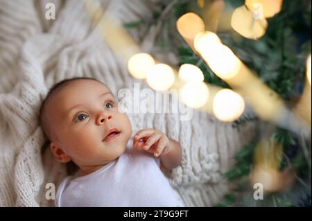 Silvester Weihnachtsfeierszene - lustiger süßer neugeborener aufgeregter Junge, der auf Strickdecke liegt und sich weihnachtliche Weihnachtsbaumgolde und Lichtergirlande anschaut. Kinderartikel-Konzept, Kinderzimmer Stockfoto