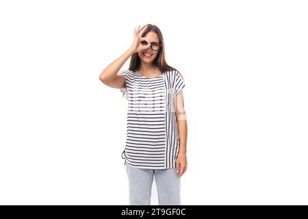 Die junge, fröhliche Brünette mit glatten Haaren in Brille trägt ein gestreiftes T-Shirt und Jeans macht eine Grimasse auf weißem Hintergrund mit Kopie Stockfoto