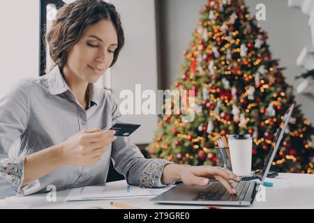 Lächelnde Frau genießt das Weihnachtsgeschäft im Internet, inmitten festlicher Einrichtung Stockfoto