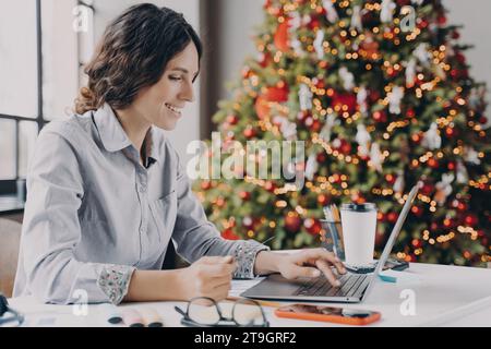 Lächelnde Frau genießt Online-Shopping auf ihrem Laptop mit einem festlichen Weihnachtsbaum im Hintergrund. Stockfoto