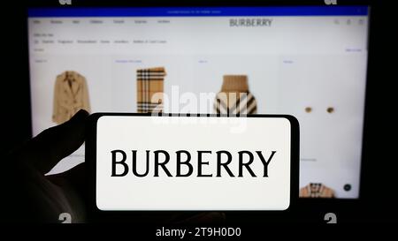 Person mit Smartphone und Logo des britischen Luxusmodeunternehmens Burberry Group plc vor der Website. Schwerpunkt auf dem Display des Telefons. Stockfoto