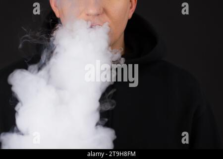 Dampfender weißer Mann, der einen Mod hält. Eine Wolke aus Dampf. Schwarzer Hintergrund. Eine elektronische Zigarette mit viel Rauch verdampfen. Vape Konzept Kopierraum auswählen Stockfoto