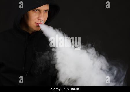 Dampfender weißer Mann, der einen Mod hält. Eine Wolke aus Dampf. Schwarzer Hintergrund. Eine elektronische Zigarette mit viel Rauch verdampfen. Vape Konzept Kopierraum auswählen Stockfoto