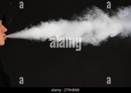 Dampfender weißer Mann, der einen Mod hält. Eine Wolke aus Dampf. Schwarzer Hintergrund. Eine elektronische Zigarette mit viel Rauch verdampfen. Vape Konzept Kopierraum auswählen Stockfoto