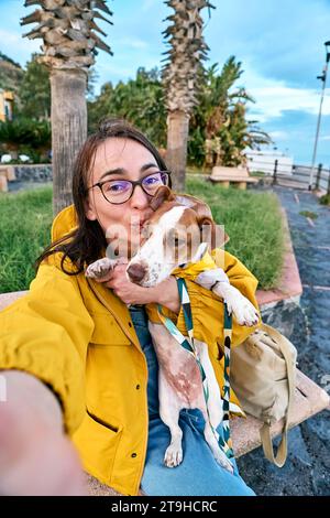 Frau umarmt ihren Jack Russell Terrier in gelbem Regenmantel und macht Selfie mit Smartphone, während sie im Frühling oder am Meer spazieren geht Stockfoto