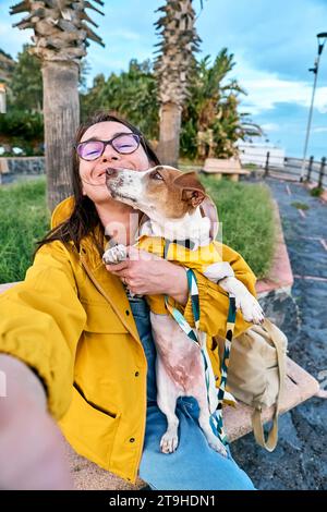 Frau umarmt ihren Jack Russell Terrier in gelbem Regenmantel und macht Selfie mit Smartphone, während sie im Frühling oder am Meer spazieren geht Stockfoto