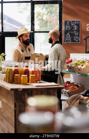 Männlicher Landwirt, der neue Charge frisch geernteter Erzeugnisse an den Lagerhalter im Nahen Osten übergibt. Kaukasier mit Hut, der dem Verkäufer mit schwarzer Schürze Kisten mit Obst und Gemüse überreicht. Stockfoto