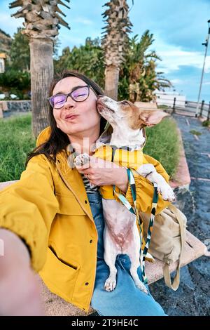 Frau umarmt ihren Jack Russell Terrier in gelbem Regenmantel und macht Selfie mit Smartphone, während sie einen Spaziergang am Meer macht Stockfoto