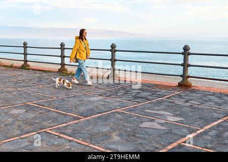 Frau in gelbem Regenmantel, die an einem Frühlings- oder Herbstnachmittag mit ihrem süßen Jack Russell Terrier entlang der Küste spaziert. Stockfoto