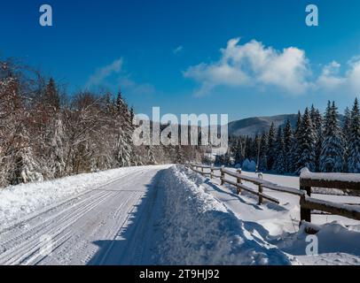 Sekundäre Landschaft alpine Straße zu abgelegenen Bergdörfern durch verschneiten Tannenwald, Schneeverwehungen und Holzzaun auf dem Weg Stockfoto