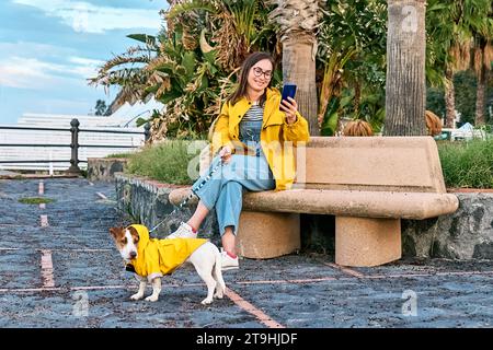 Lächelnde Frau in gelber Jacke mit Smartphone gekleidet, während sie mit ihrem süßen Jack Russell Terrier in einem gelben rai einen Spaziergang am Meer macht Stockfoto