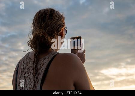 La Union, La Union, El Salvador - 30. November 2022: Erwachsene Frau in einem ärmellosen Shirt hält ein Smartphone, um ein Foto des Sonnenuntergangs auf einem Boot PR zu machen Stockfoto