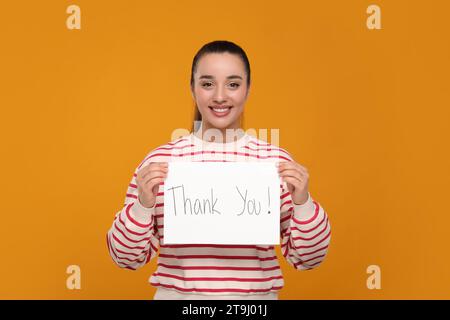 Glückliche Frau, die Karte mit Phrase hält Danke auf orangem Hintergrund Stockfoto