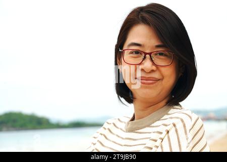 Porträt einer asiatischen Frau mittleren Alters mit Brille, lächelnd, Nahaufnahme Porträt, draußen mit Himmel und Meer Hintergrund. Stockfoto