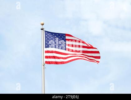 Amerikanische Flagge auf silbernem Metallfahnenmast, der im Wind weht, blauer bewölkter Himmel im Hintergrund. Stockfoto