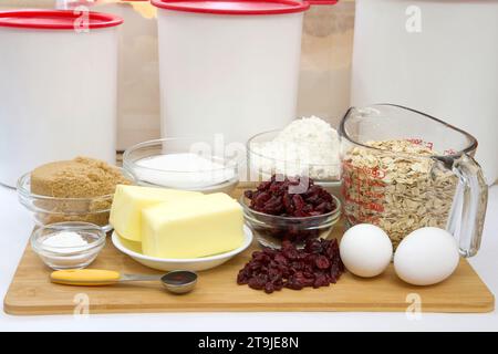 Gemessene Zutaten in Schüsseln und Bechern für Cranberry Haferflockenkekse. Auf Holzschneidebrett mit Pergamentpapier darunter. Kunststoffkanister im Backg Stockfoto