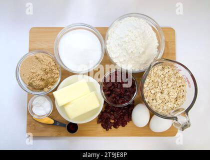 Flache Anordnung der abgemessenen Zutaten in Schüsseln und Bechern für Cranberry-Haferflockenkekse. Auf Holzschneidebrett mit Pergamentpapier darunter. Stockfoto