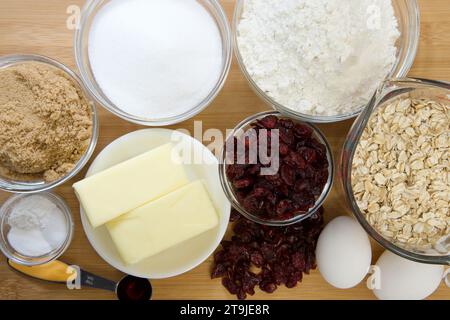 Flache Anordnung der abgemessenen Zutaten in Schüsseln und Bechern für Cranberry-Haferflockenkekse. Auf Holzschneidebrett mit Pergamentpapier darunter. Stockfoto