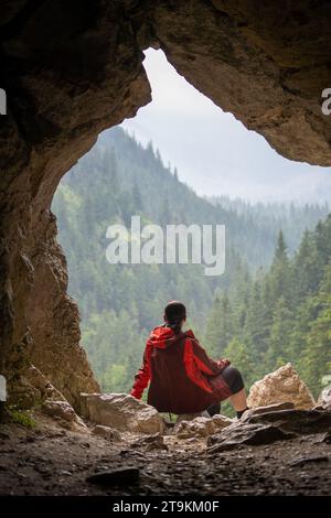 Eine Frau sitzt an einer Höhle und schaut auf die Berglandschaft, Blick vom Inneren der Höhle im Sommer Stockfoto