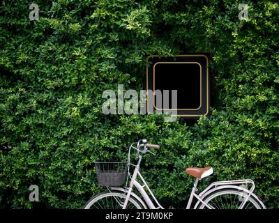 Quadratisches schwarzes leeres Ladenschild, umgeben von grünem Busch mit einem Vintage-Fahrrad. Mockup der gelben Rahmenbeschilderung vor Restaurant, Geschäft oder Café, mou Stockfoto