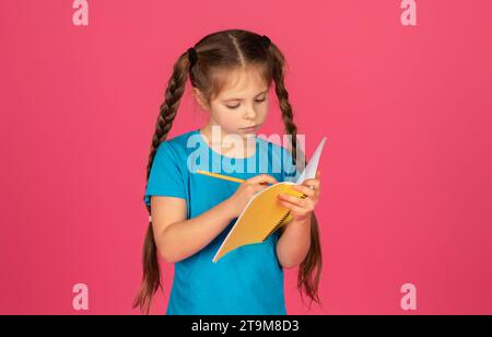 Ein fokussiertes kleines Mädchen schreibt in Notizbuch, während es auf rosa Hintergrund steht Stockfoto