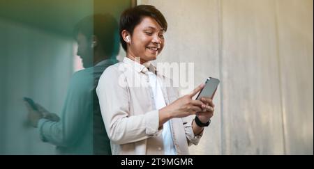 Junger asiatischer Mann, der draußen steht, Smartphone und Ohrhörer benutzt Stockfoto