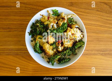 Vegetarische Mahlzeit mit im Ofen geröstetem Blumenkohl, Grünkohl und roter Paprika über braunem Reis in einer weißen Schüssel auf einem Holztisch. Stockfoto