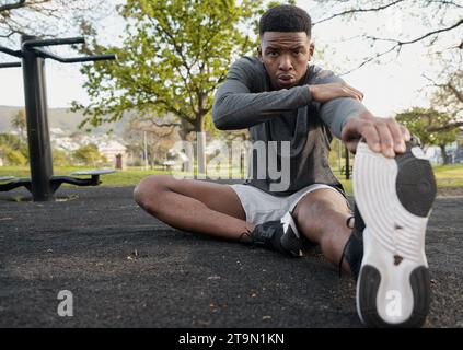 Junger schwarzer Mann in Sportbekleidung sitzend und ausatmend, während er sich im Park dehnt Stockfoto