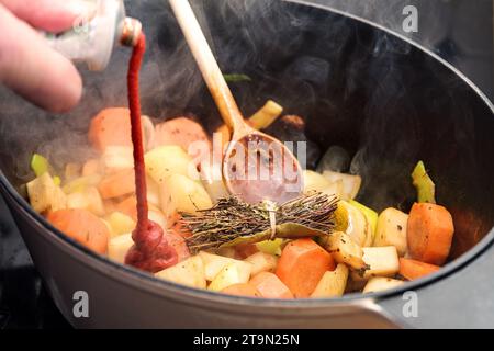 Tomatenpaste wird in einen Topf mit dampfendem geröstetem Gemüse wie Karotte, Zwiebeln, Sellerie, Lauch und einem Kräuterbouquet gegossen Stockfoto