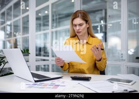 Junge Buchhalterin, Finanzanalystin, die mit Dokumenten im Büro, Problemen mit Konten und Berichten arbeitet, sieht besorgt aus und spreizt die Hände. Stockfoto