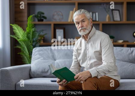 Porträt eines grauhaarigen älteren Mannes, der zu Hause auf der Couch sitzt, ein Buch in der Hand hält und ernsthaft in die Kamera schaut. Stockfoto