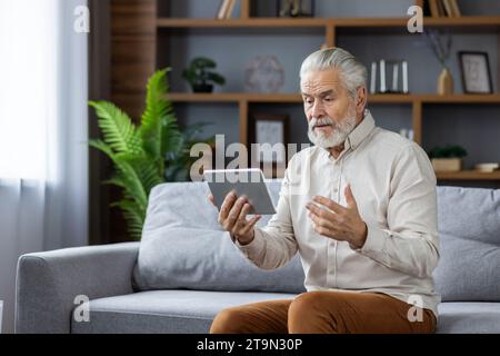 Senior grauhaariger Mann kommuniziert per Videoanruf von einem Tablet mit einem Arzt, führt eine Online-Konsultation durch, erklärt das Problem, sitzt zu Hause auf dem Sofa. Stockfoto