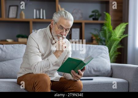 Ein älterer grauhaariger Mann sitzt auf der Couch zu Hause in einer Schürze und liest ein interessantes Buch mit Konzentration. Stockfoto