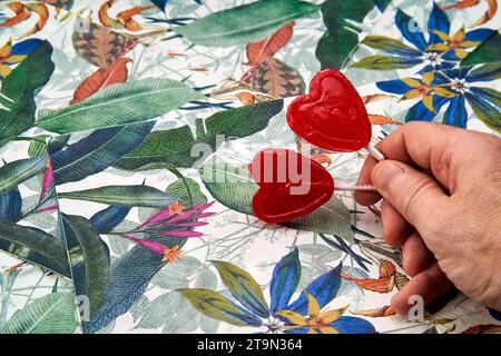 Die Hand des Mannes hält zwei rote Herzlutscher mit tropischem Hintergrund. Stockfoto