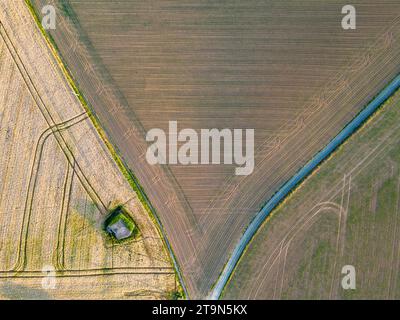 Dieses Bild von oben fängt die geometrische Schönheit des Ackerlandes ein, mit seinen verschiedenen Braun- und Grüntönen, die einen Flickenteppich aus landwirtschaftlichem Boden bilden. Ein geschwungener blauer Wasserweg zieht sich durch die Landschaft und bildet einen starken Kontrast zu den geraden Linien der Kulturfelder. Ein einsames Gebäude, möglicherweise ein Nebengebäude, befindet sich am Rande eines Feldes und bietet ein Gefühl von Größe und menschlicher Präsenz in diesem riesigen ländlichen Wandteppich. Ländliches Flickwerk: Luftaufnahme von Farmland und Wasserweg. Hochwertige Fotos Stockfoto