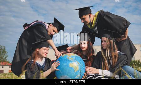 Absolventen in schwarzen Gewändern untersuchen einen geografischen Globus, der auf dem Gras sitzt. Stockfoto
