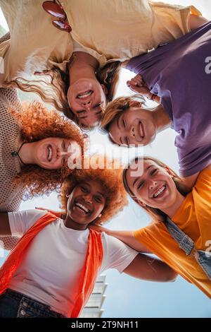 Vertikale Tiefwinkelansicht von fünf Mädchen im Teenageralter, die lachen und in die Kamera schauen. Eine Gruppe junger Frauen, die lächelnd nach unten schauen. Echte sorglose Damen, die sich zusammen umarmen. Begeisterte Freunde posieren für ein Porträt. Hochwertige Fotos Stockfoto