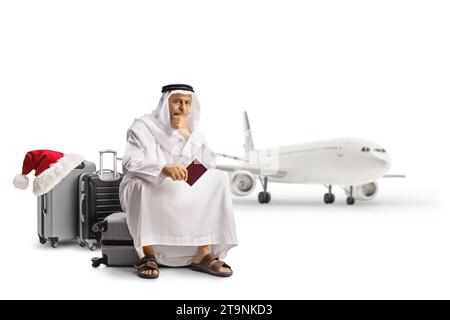 Besorgter arabischer Mann, der auf einem Koffer sitzt und einen Pass hält und auf einen Flug auf weihnachten wartet, isoliert auf weißem Hintergrund Stockfoto