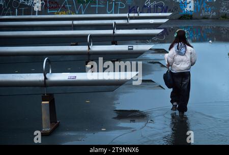 Berlin, Deutschland. November 2023. 19.11.2023, Berlin. Ein Mädchen mit Kopfhörern geht nach einem Regenguss an einem nassen Herbsttag in der Abenddämmerung durch eine große Pfütze rund 100 Meter vom Potsdamer Platz im Tilla-Durieux-Park. Die junge Frau trägt Gummistiefel zu ihrer modernen Kleidung und wird deshalb nicht nass. Kredit: Wolfram Steinberg/dpa Kredit: Wolfram Steinberg/dpa/Alamy Live News Stockfoto