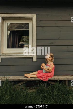 Porträt eines Mädchens, das auf einer Bank neben dem Haus sitzt und eine Puppe umarmt. Stockfoto