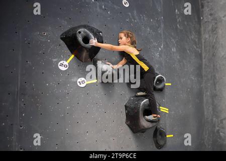 Das kleine Mädchen trainierte klettert in der Kletterhalle an der Wand. Kind mit braunem Haar in Sportkleidung, das ohne Hilfe im Boulderzentrum für Kinder arbeitet. Das tapfere und starke Mädchen klettert auf die falsche Wand Stockfoto