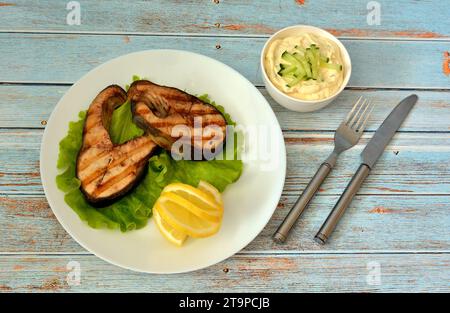 Zwei rosafarbene Lachssteaks auf einem Salatblatt mit Zitronenscheiben auf einem Holztisch, neben einer Tasse mit Sauce und Besteck. Draufsicht, flach. Stockfoto