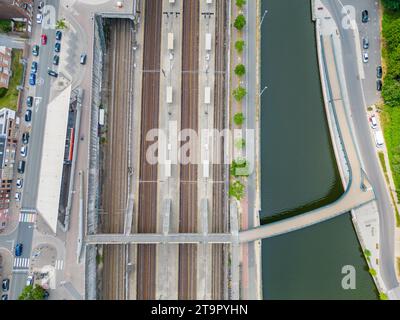 Das Bild zeigt eine Ansicht von oben nach unten eines Bahnhofs neben einem kurvenreichen Fluss, der den Zusammenfluss verschiedener Transitarten veranschaulicht. Die Bahngleise verlaufen parallel, von Bahnsteigen unterbrochen, und sind bereit für die dazugehörigen Wagen. Der Fluss, der von Promenaden begrenzt wird, reflektiert die umgebenden Strukturen und fügt einen fließenden Kontrast zu den starren Linien der Eisenbahn hinzu. Diese städtische Momentaufnahme erfasst das organisierte Chaos des Verkehrs in Bewegung und hebt die Infrastruktur hervor, die den Rhythmus des Stadtlebens unterstützt. Zusammenfluss von Transit: Bahn und Fluss. Hochwertige Fotos Stockfoto