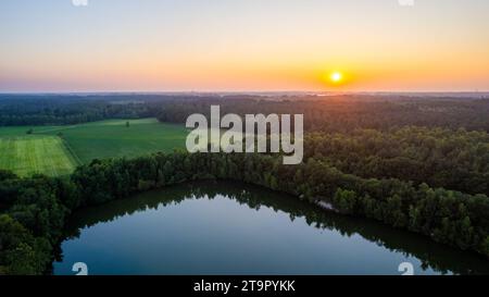 Dieses Bild ist eine heitere Darstellung der Dämmerung, während das letzte Licht des Tages einen abgelegenen See schmückt. Die Sonne, eine feurige Kugel, schwebt am Horizont und wirft ein Spektrum warmer Farben über den Himmel. Der umliegende Wald steht wie eine dunkle Silhouette vor dem leuchtenden Himmel, während der See darunter das schwindende Licht reflektiert. Der Kontrast zwischen den dunkleren Wäldern und dem beleuchteten Himmel erzeugt einen Moment der natürlichen Pracht und fängt den friedlichen Übergang von Tag zu Nacht ein. Die Abenddämmerung steigt auf einem ruhigen See ab. Hochwertige Fotos Stockfoto