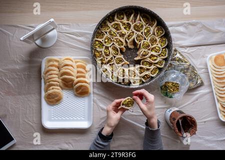 Hände halten Qatayef gefüllt mit Schokolade und mit Pistazien belegt auf einem Holztisch, mit einem Teller, der später im Ofen als Ramadan swe zubereitet wird Stockfoto